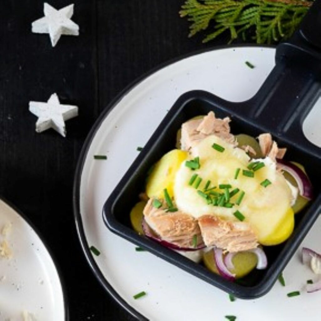 Raclette Pfännchen mit Kartoffeln und Thunfisch wird auf einem weißen Teller mit Schnittlauch gezeigt.