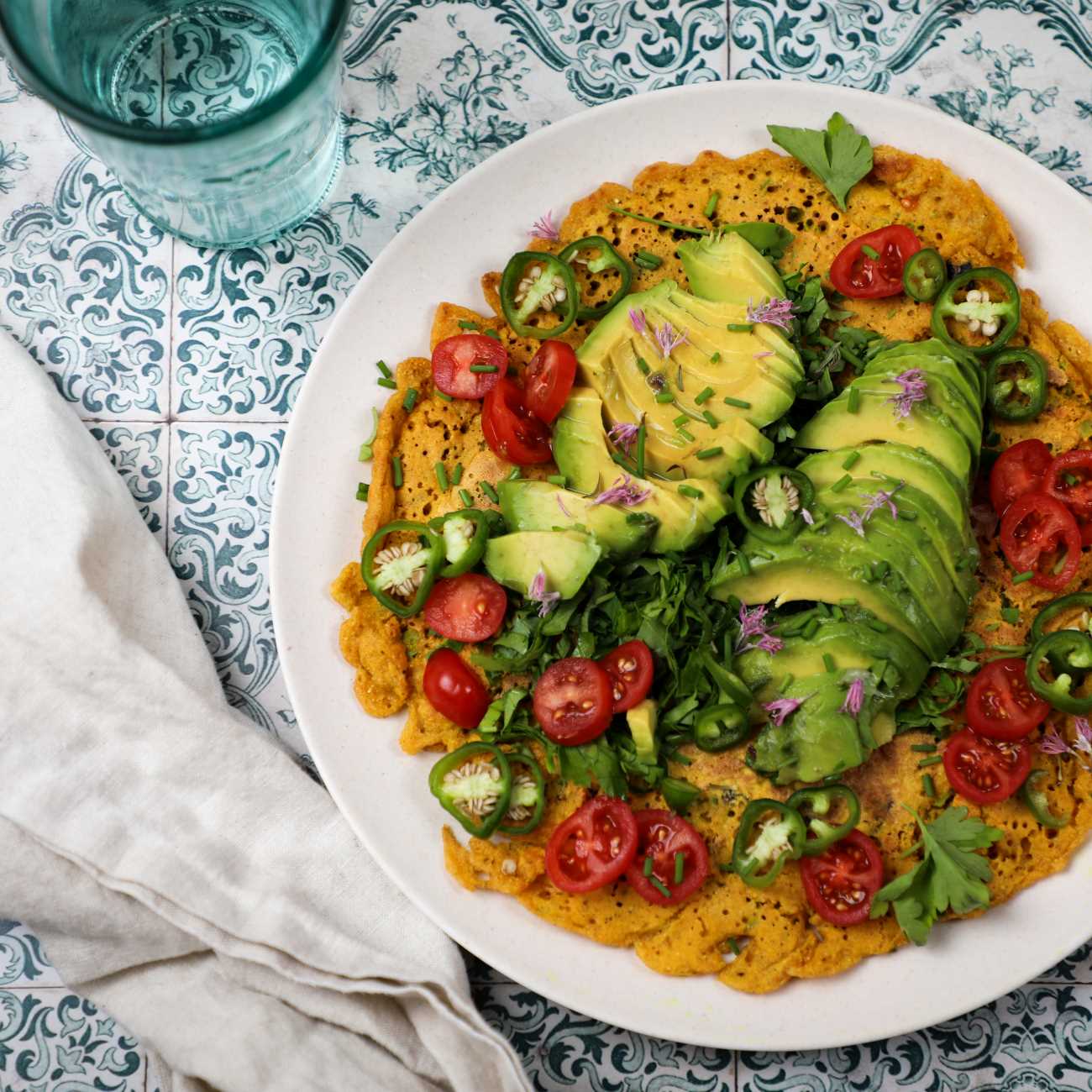 Veganes Omelett wird mit Salat und Avocado auf einem Teller auf bunten Fliesen gezeigt.