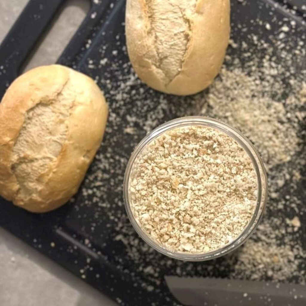Semmelbrösel selber machen: Semmelbrösel werden in einem Glas auf einem Schneidebrett mit Brötchen gezeigt.