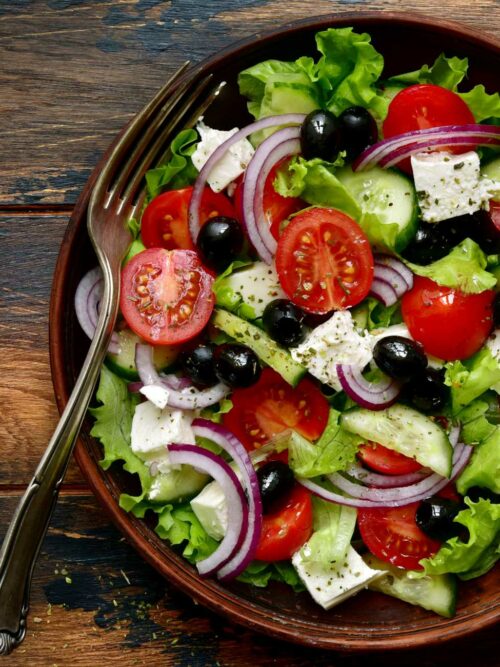 Griechischer Salat wird in einer braunen Schüssel auf Holzuntergrund gezeigt.
